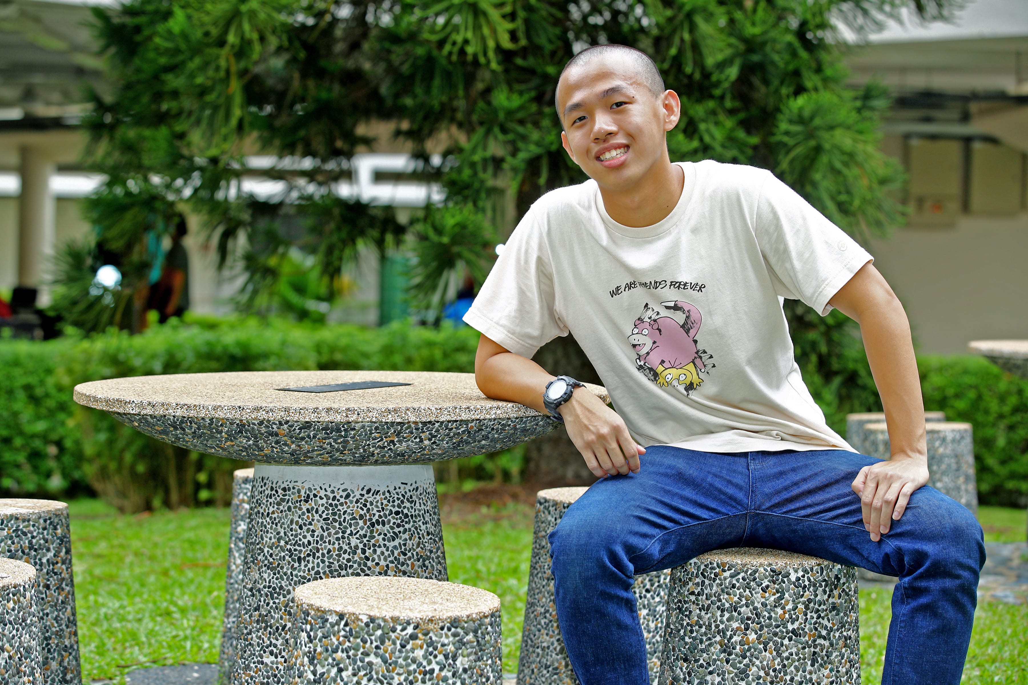 Smiling person in a white T-shirt and jeans sits on a stone stool beside a round stone table outdoors.